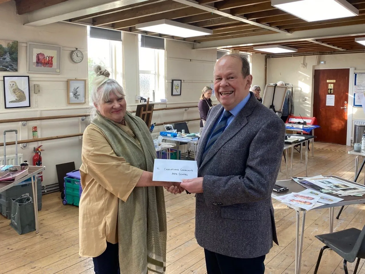 A man and woman smile whilst shaking hands and holding a cheque inside a community arts centre studio.