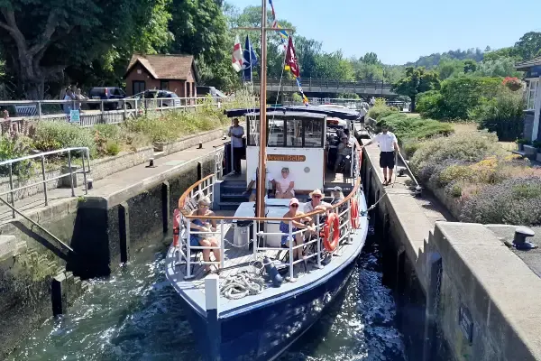 A passenger riverboat with visitors on deck navigates through a canal lock on a sunny day.