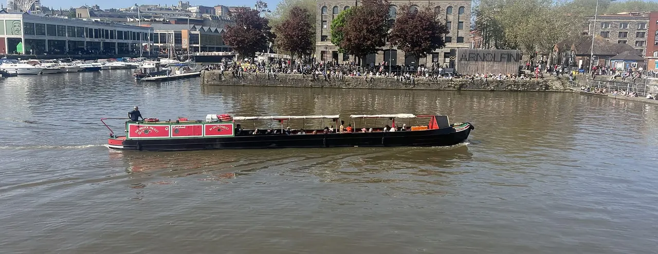 A narrowboat with red and black livery carries passengers along a river past a crowded waterfront lined with historic buildings.