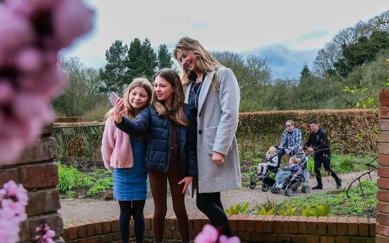 Woman and two children standing in a walled garden with pink flowering shrubs, other visitors visible in background.