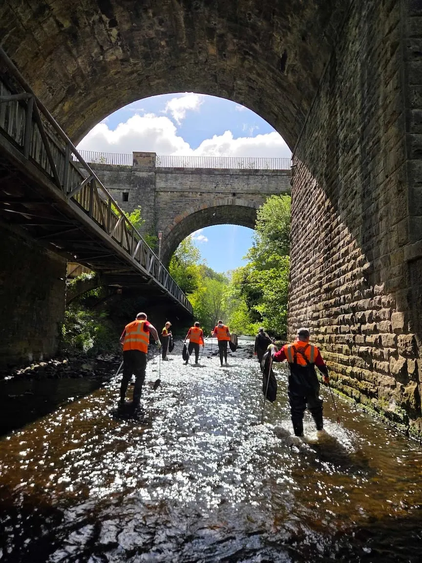 Group of people in orange safety vests walking through shallow water beneath historic stone railway viaducts and arches.
