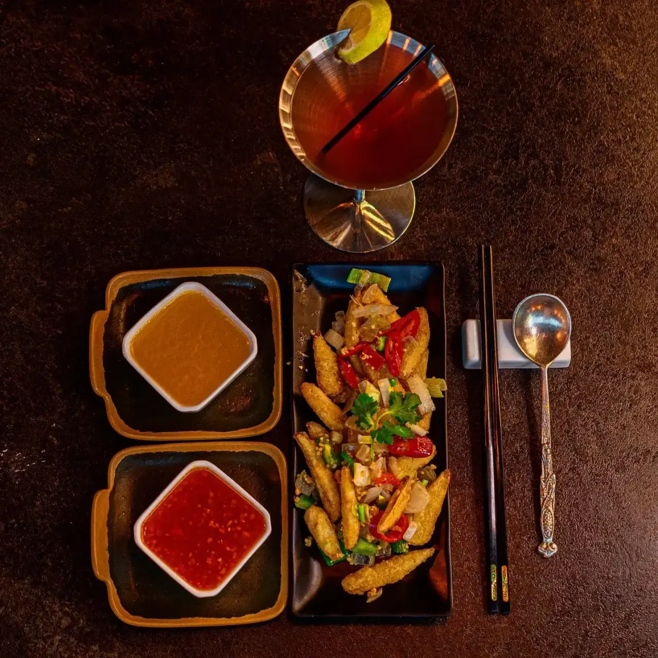 Overhead view of Asian meal with fried wontons, dipping sauces, chopsticks, and a cocktail with yellow garnish on dark surface.
