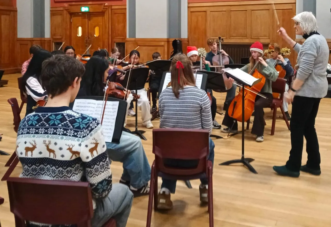 Young musicians and children in festive Santa hats rehearse with string instruments under a conductor in a wooden concert hall.