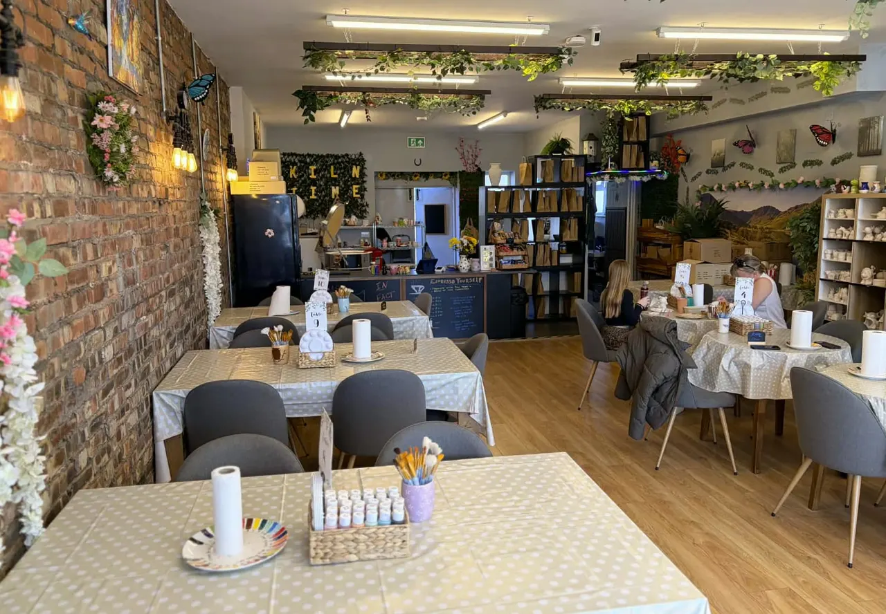 Interior of a pottery studio with tables set up for creative activities, shelving displaying ceramic pieces, and visitors seated at workstations.