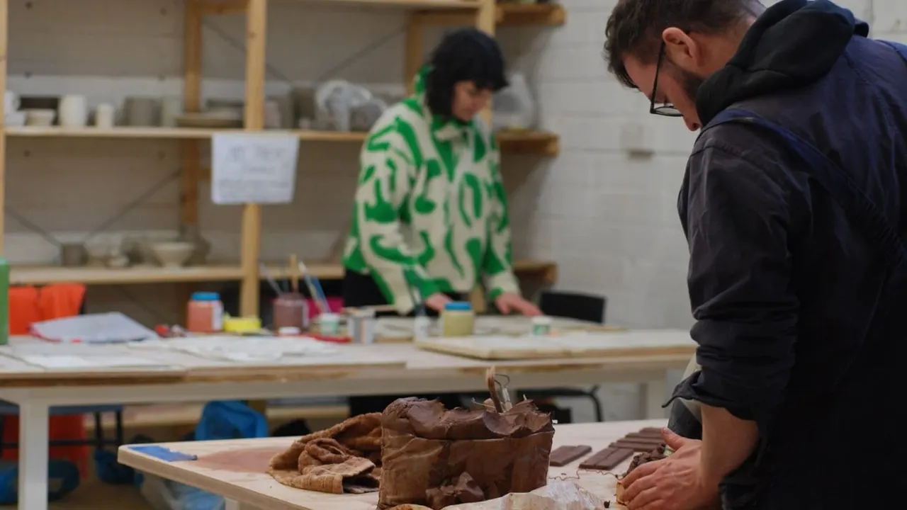 Two visitors participate in a clay pottery workshop, with one shaping clay at a table whilst an instructor in a green patterned top supervises nearby.