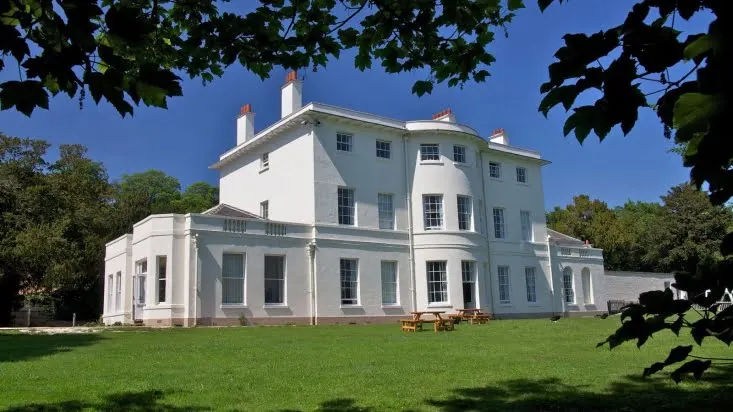 Large white Georgian mansion set in expansive green grounds, framed by tree foliage.