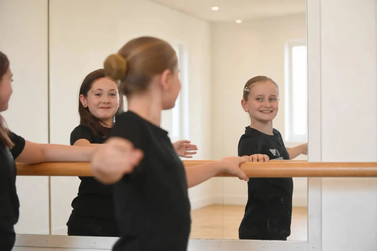 Young children in black dance attire at a ballet barre during a class, reflected in a mirror.