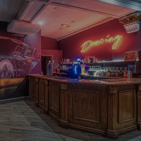 Bar counter with neon 'Dancing' sign and bottles lit by red and blue lighting in an empty venue.