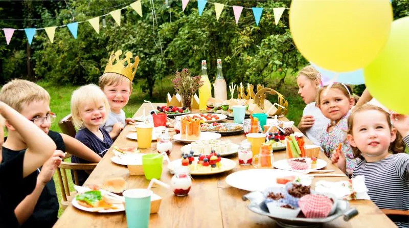 Children seated at a long outdoor table for a garden party with food, drinks, bunting and balloons.