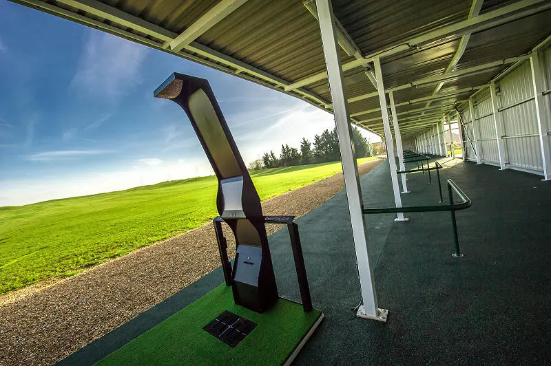 Golf ball launcher on putting green under covered bay, with countryside visible beyond.