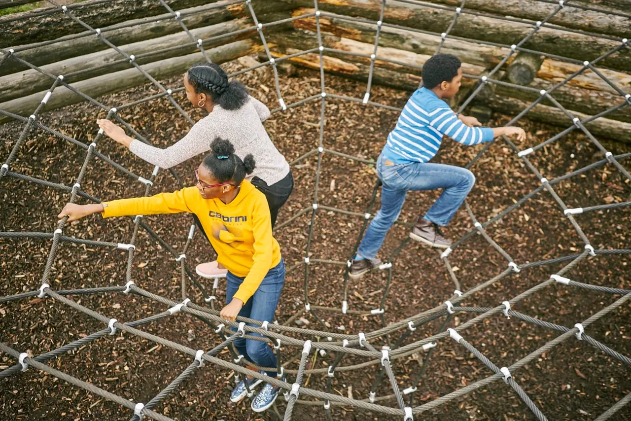 Three children balancing on a rope obstacle course made of wooden logs and cable netting.