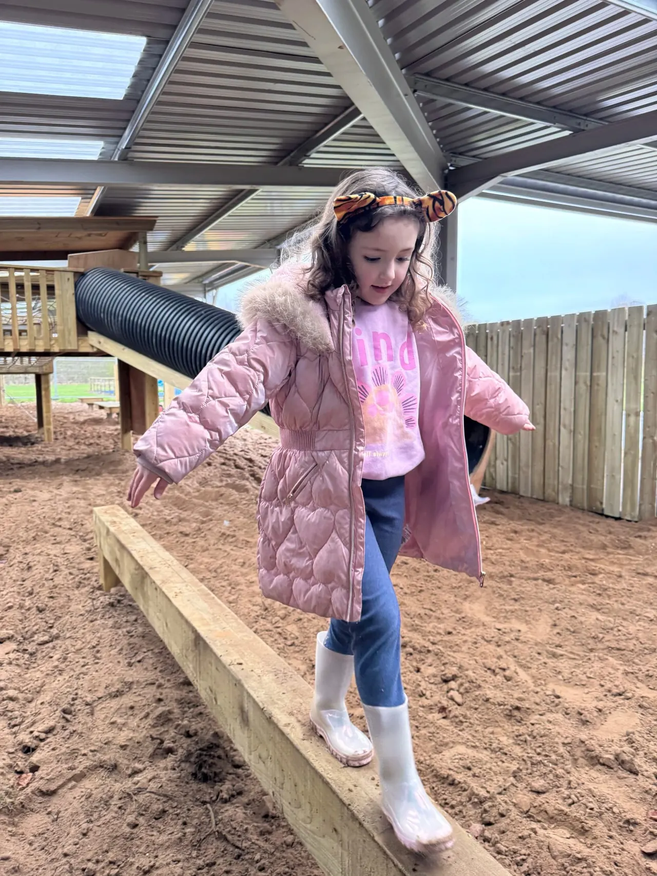 A young girl balances on a wooden beam in a farm play area with covered structures and fenced enclosures behind her.