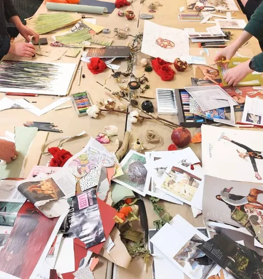 Two people create a floral-themed artwork at a table scattered with sketches, dried flowers, and art supplies.