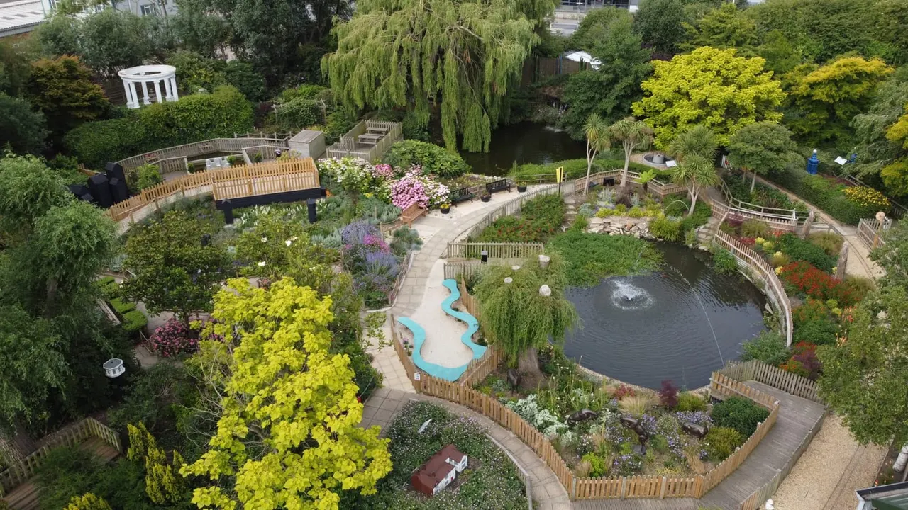 Aerial view of a landscaped garden with a pond, winding pathways, wooden fencing, and colourful planted areas including yellow and pink flowering shrubs.