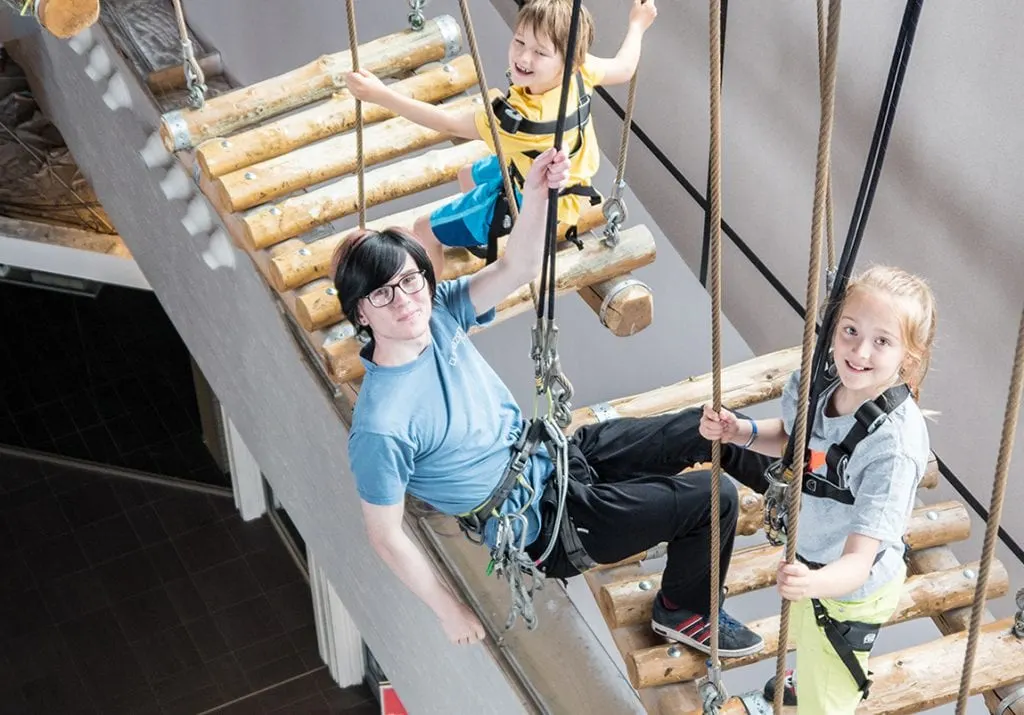 An adult and three children navigate a rope and log climbing course indoors, wearing safety harnesses and smiling.