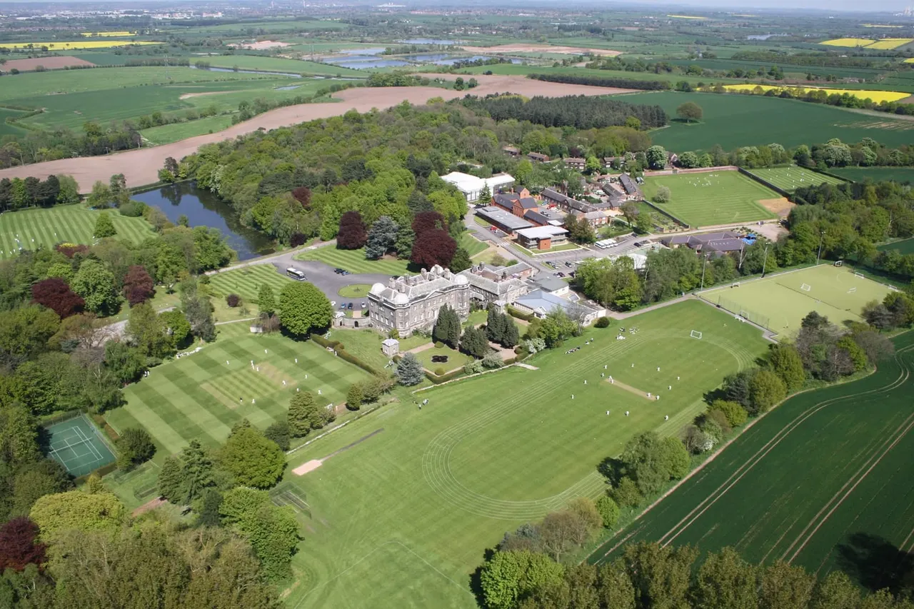 Aerial view of a country estate with manicured grounds, sports fields, a historic stone building, and surrounding parkland and farmland.