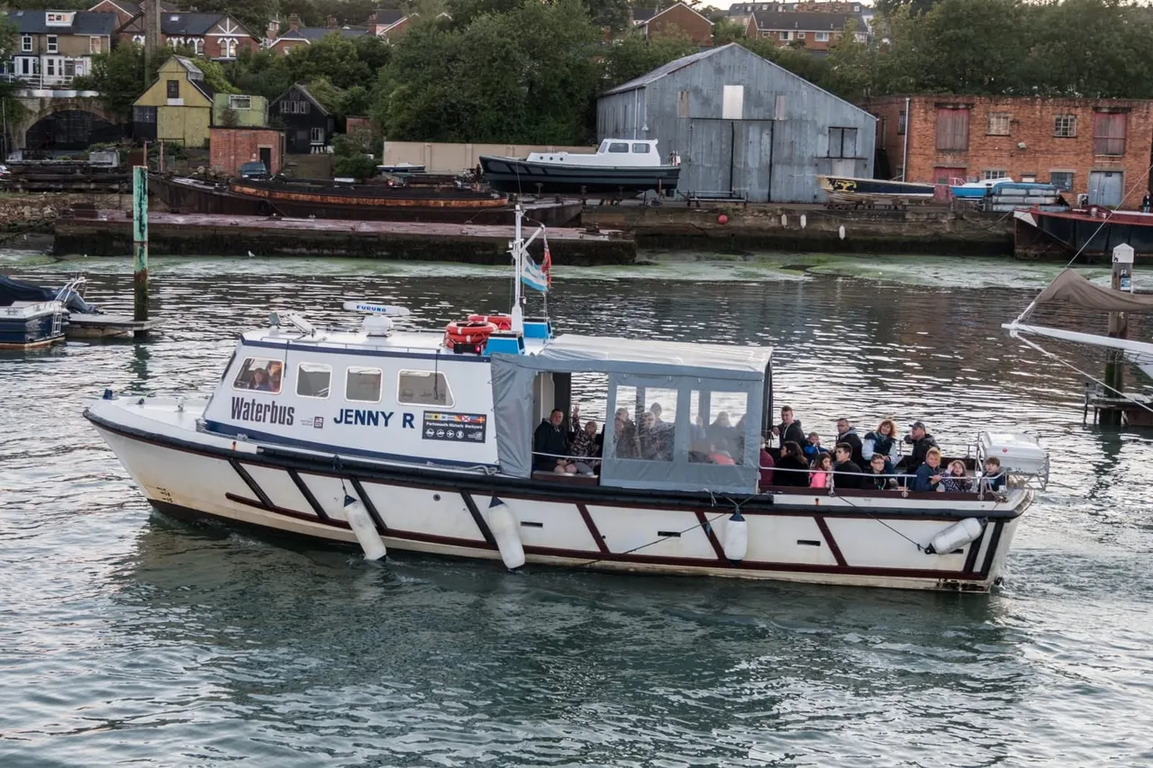Waterbus Jenny R carrying passengers on a river cruise past historic industrial waterfront buildings and moored boats.