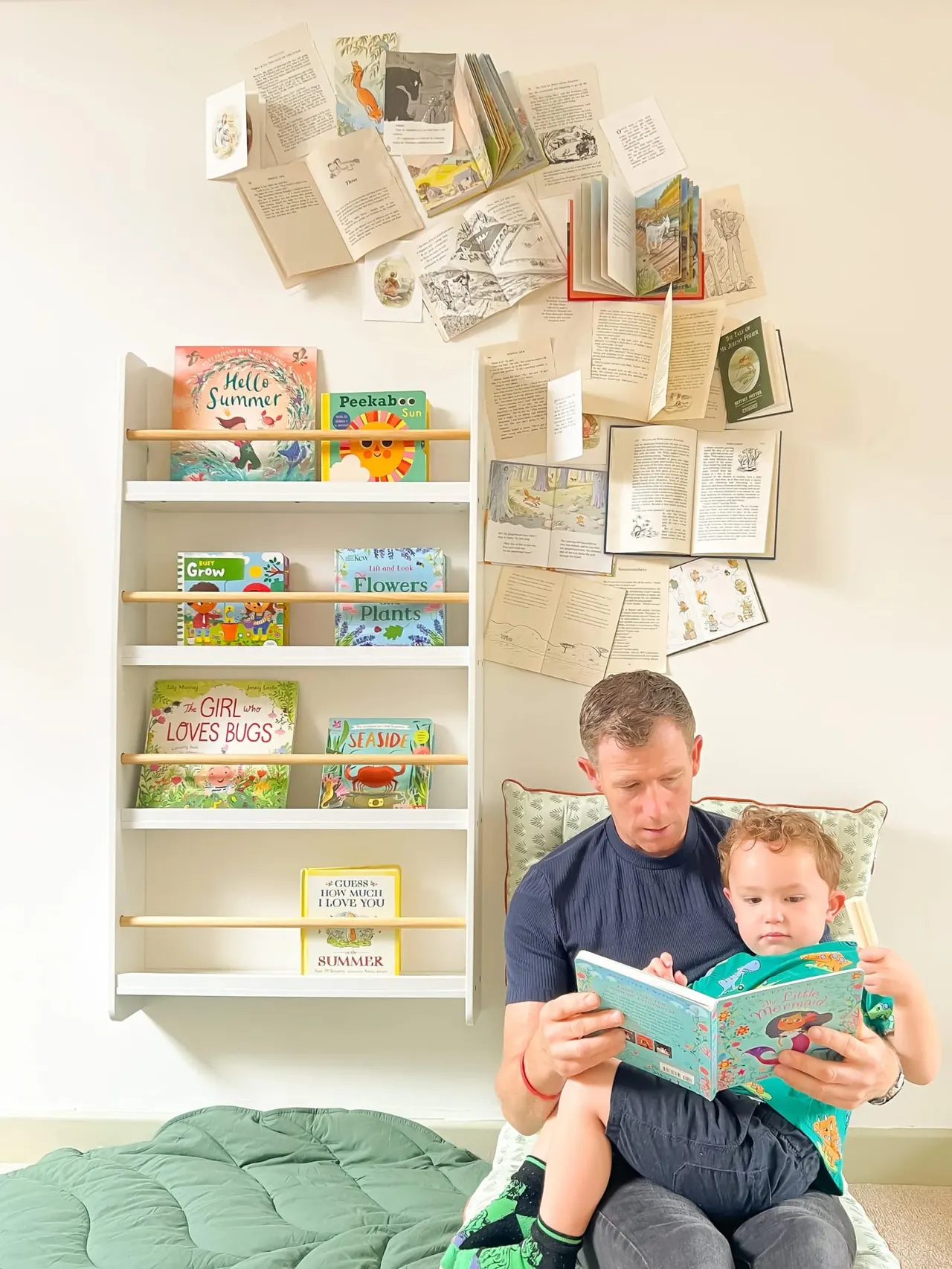 Adult and young child reading a picture book together, sat in front of a shelving unit displaying children's books and open pages on the wall.