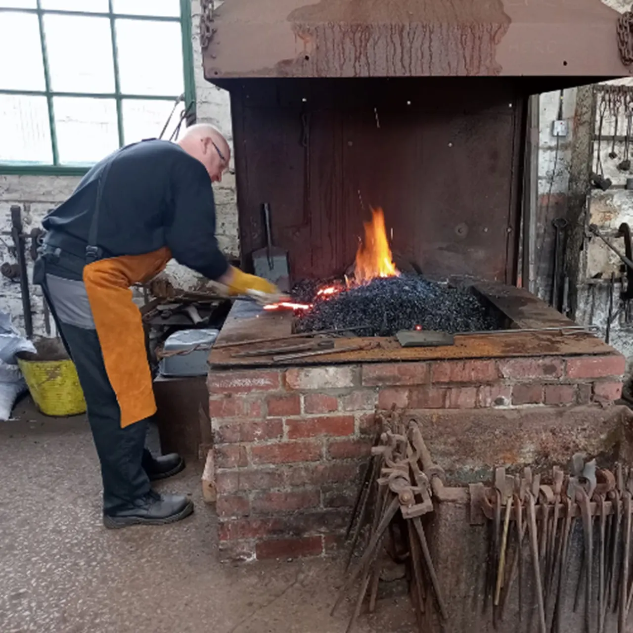 A blacksmith tends to a forge with glowing coals and flames, using a hammer and tongs to work metal at an outdoor heritage site.