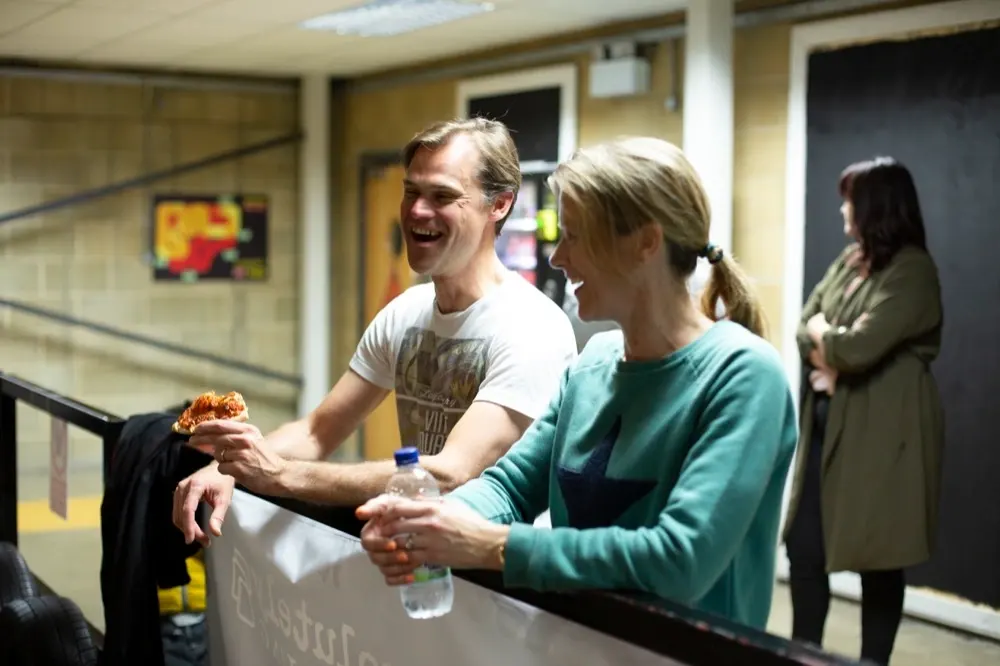 A man and woman smile whilst sharing food at an indoor leisure venue with a basketball court visible in the background.