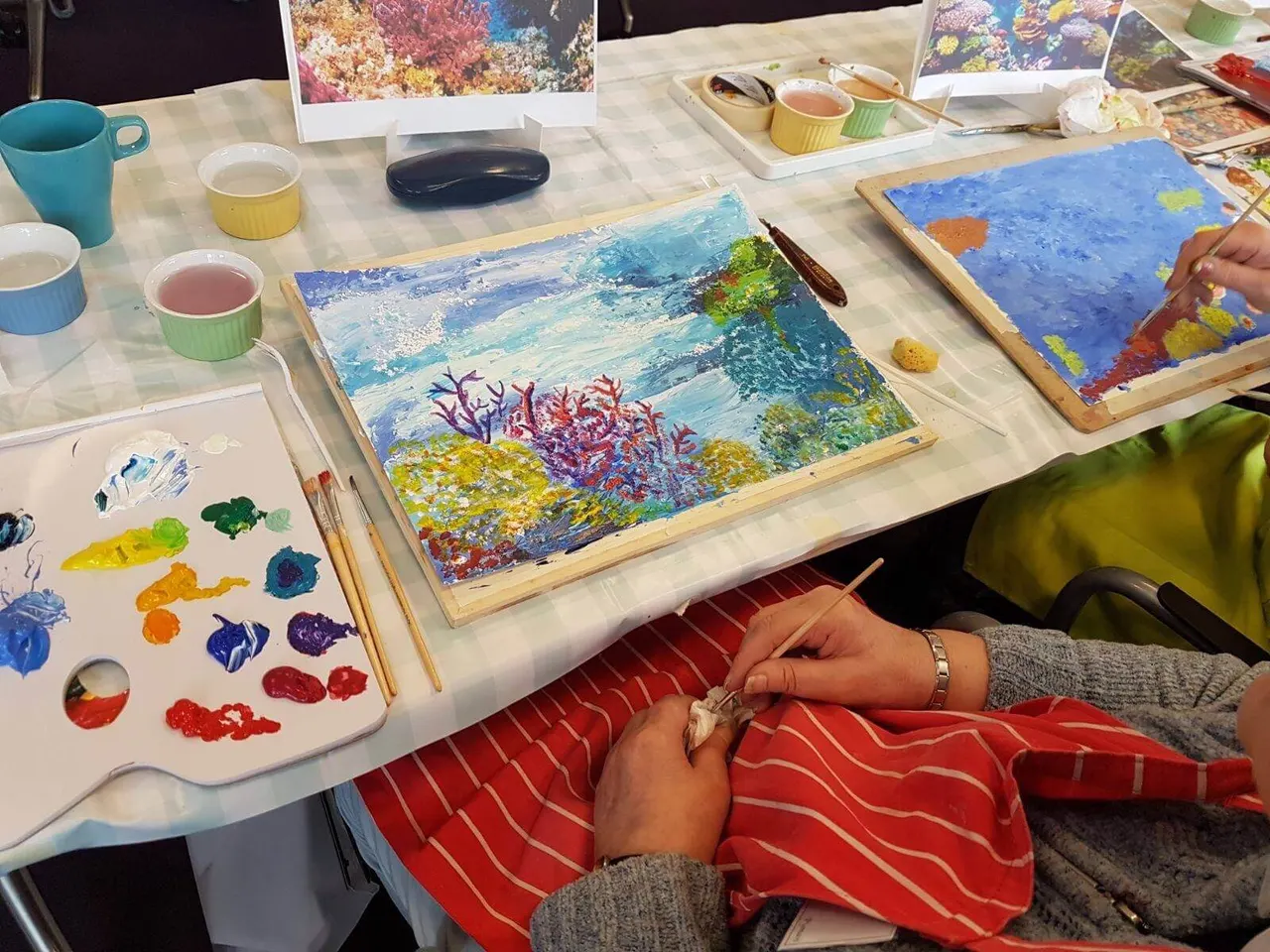 Person painting ocean and coral reef scenes at an art class workshop table with supplies.