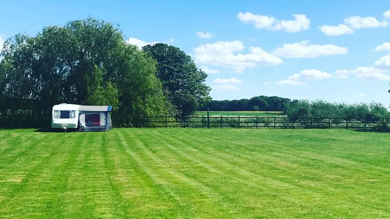 Caravan parked in open grassland with mature trees and fenced pasture under blue sky.