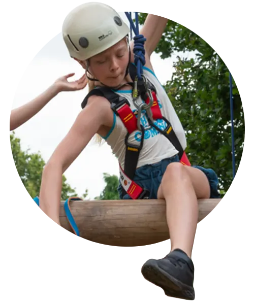 Child in safety helmet and harness climbing a wooden obstacle during an adventure course activity.