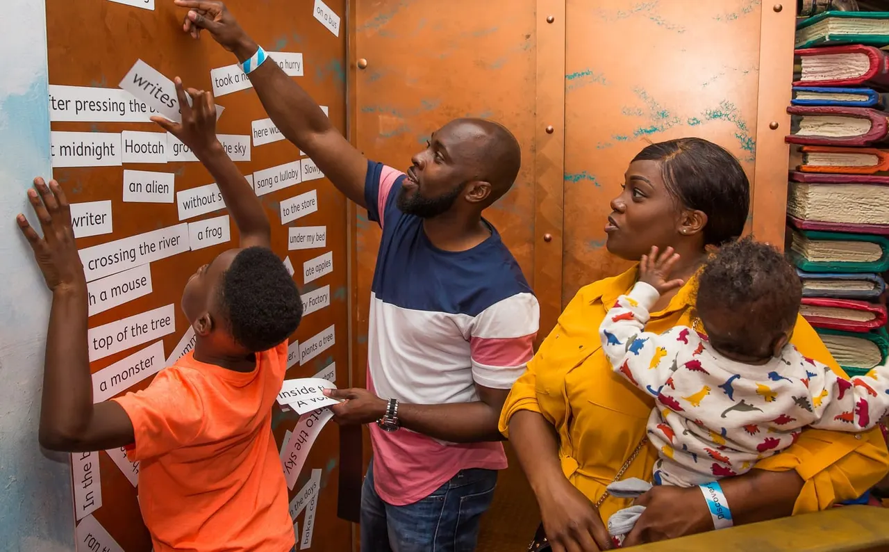 Two adults and two children interact with a word-card display on an orange wall during a literacy activity.
