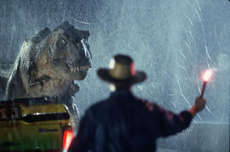 This is a GRAPHIC - it appears to be a scene from a film or designed promotional image overlaid with text effects, showing a person with a torch facing a large dinosaur head in heavy rain.