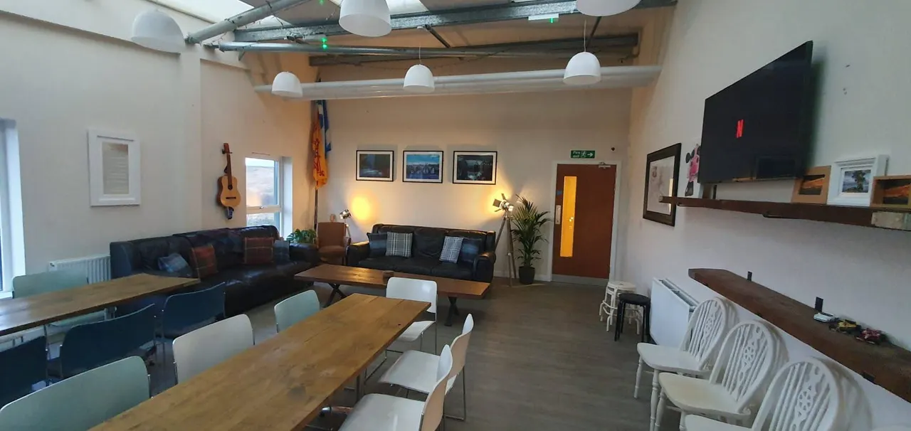 Spacious communal lounge and dining area with wooden tables, black sofas, a guitar on the wall, and a television.