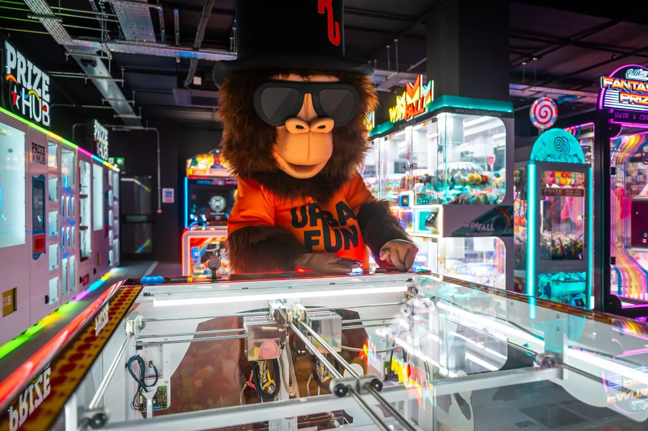 Oversized ape mascot wearing sunglasses and orange shirt stands behind arcade claw machine in colourful arcade venue with neon signs.