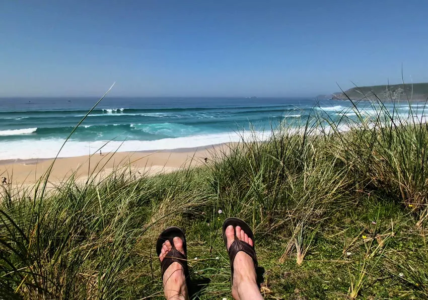 Person's feet in sandals resting on coastal grass overlooking a sandy beach with turquoise waves and cliffs.