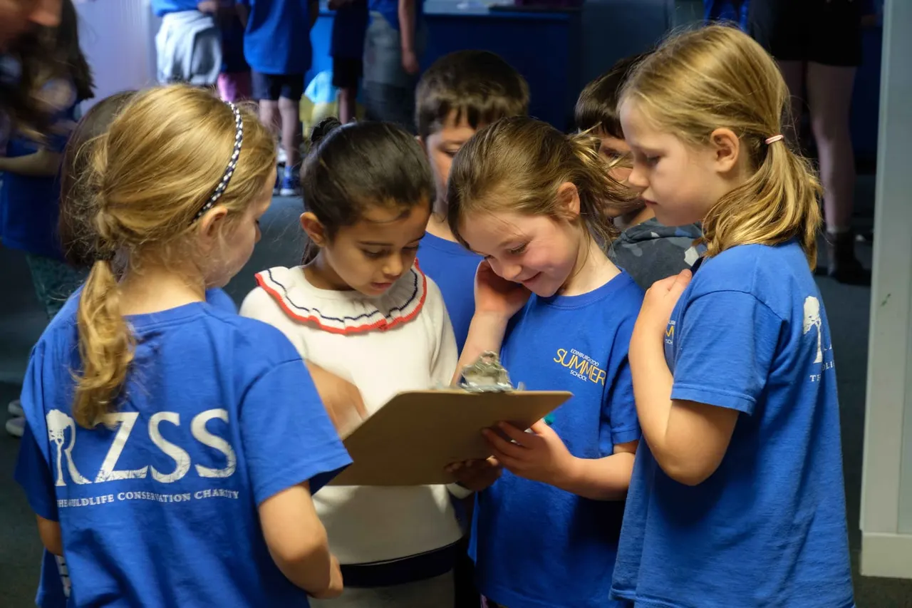 Group of children wearing blue zoo volunteer shirts looking at a tablet together indoors.