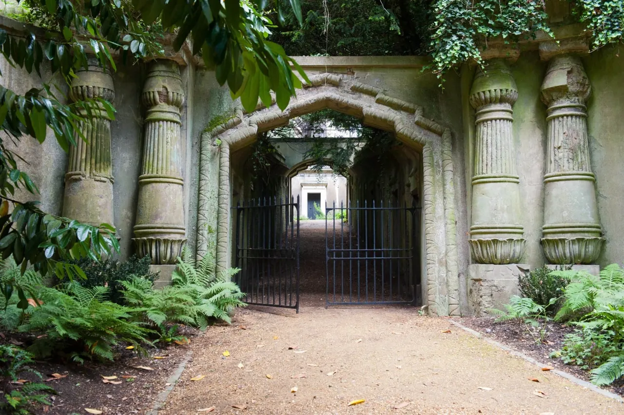Stone archway with ornamental columns and iron gates, surrounded by lush green foliage and climbing plants.