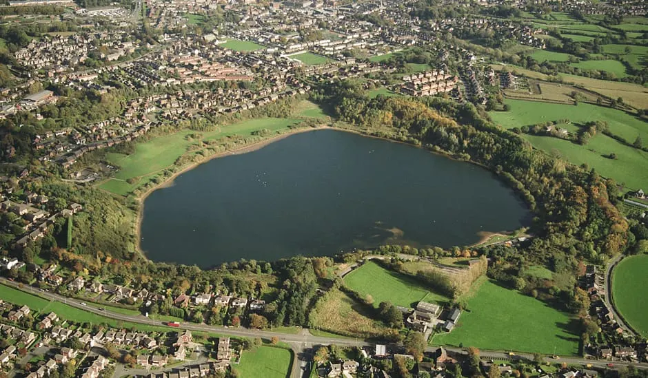 Aerial view of a large lake surrounded by woodland and green fields, with suburban housing visible around its perimeter.