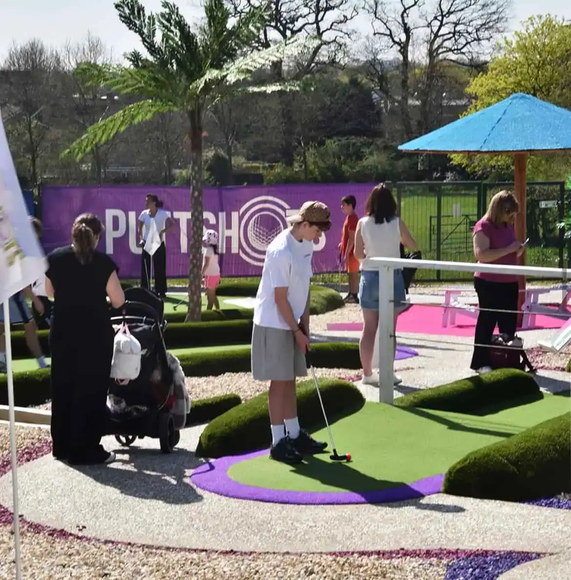 Adults and children playing mini golf on a colourful purple and green course with a blue parasol, surrounded by trees.