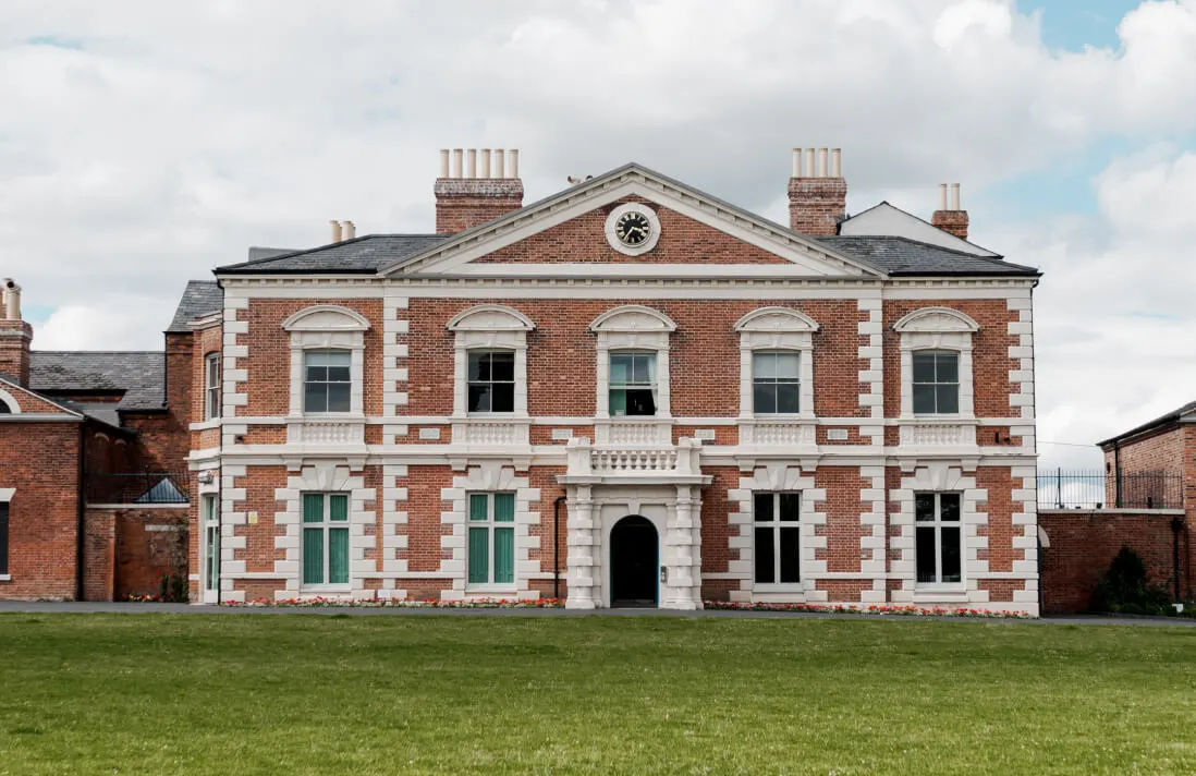 Georgian brick mansion with clock tower and manicured lawn in front, surrounded by cloudy sky.