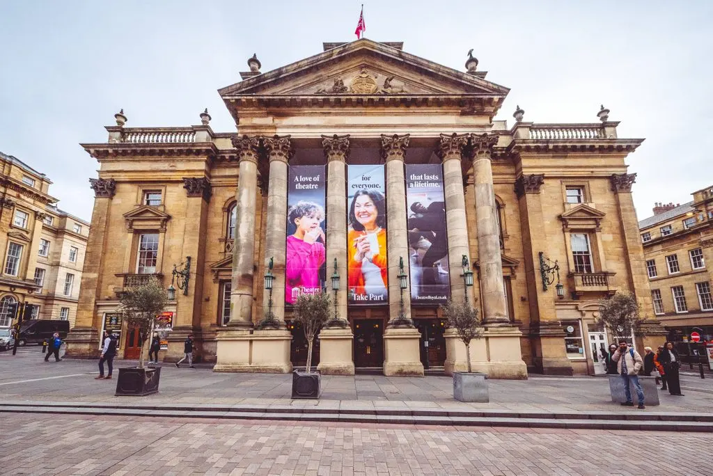 Grand neoclassical theatre building with columned façade and promotional banners, people walking in front on paved square.