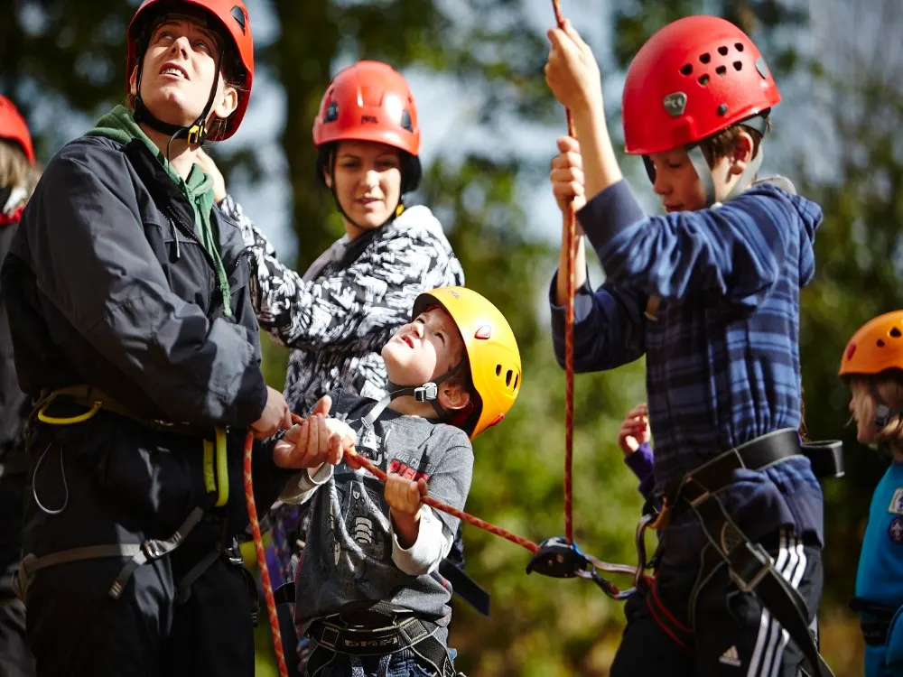 Children and adults wearing coloured helmets practising rock climbing with ropes outdoors.