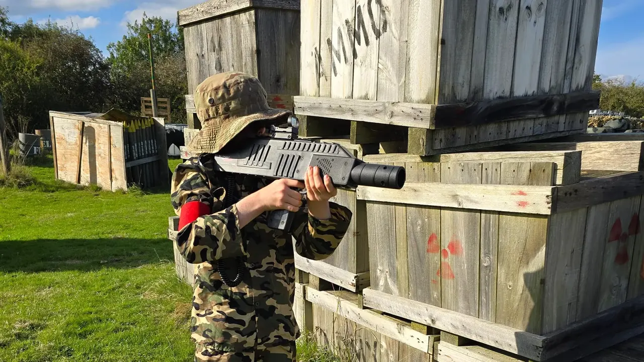 Person in camouflage gear aiming a laser tag gun at wooden barriers during an outdoor game.
