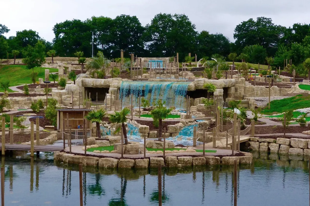 A themed water play area with stone ruins, a blue waterfall feature, palm trees, and climbing structures surrounding a lagoon.