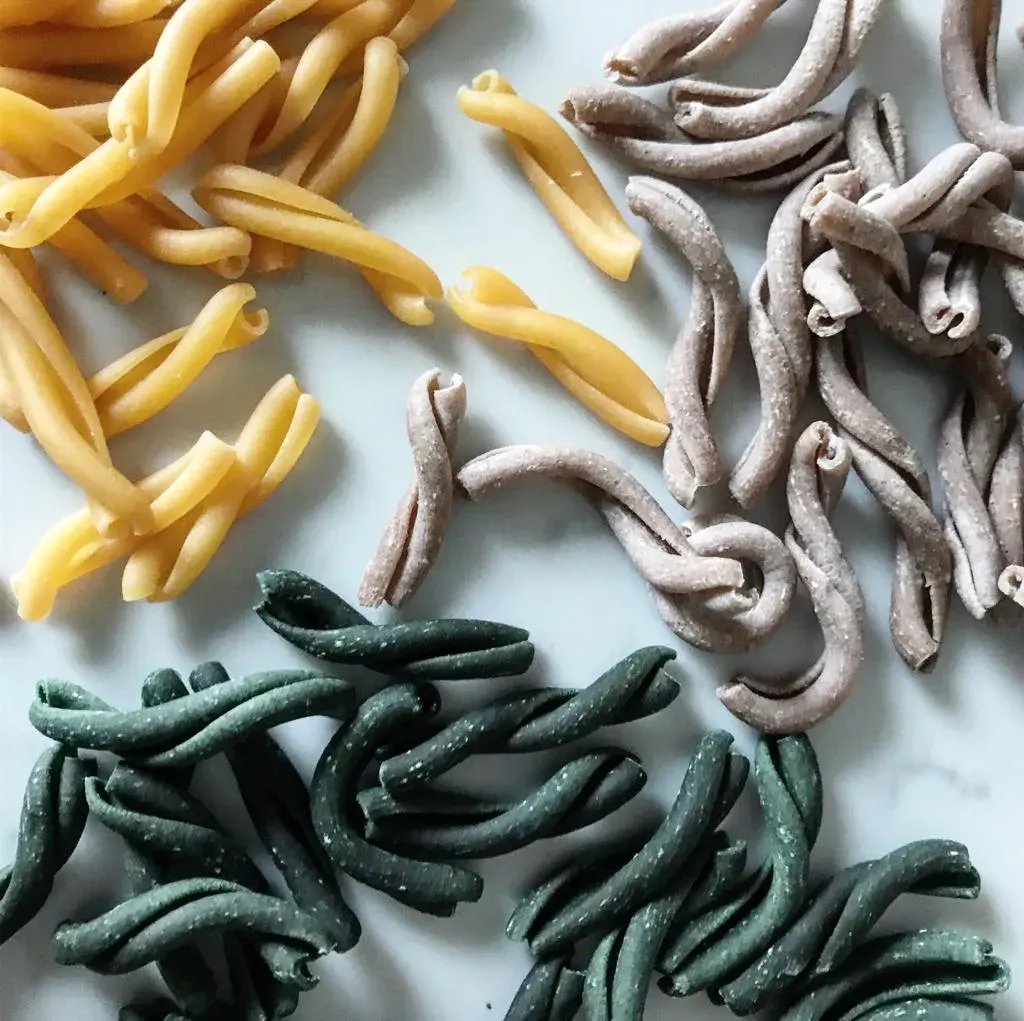 Flat lay of three varieties of uncooked pasta: yellow egg tagliatelle, brown wholemeal pappardelle, and green spinach tagliatelle on white surface.
