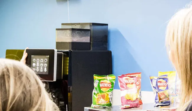 Two people with blonde hair viewing a vending machine stocked with snack packets against a blue wall.