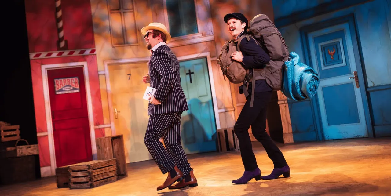 Two actors perform a comedic scene on stage with colourful set pieces depicting storefronts and doors.