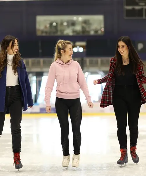 Three young people ice skating together indoors, arms linked, wearing casual winter clothing.