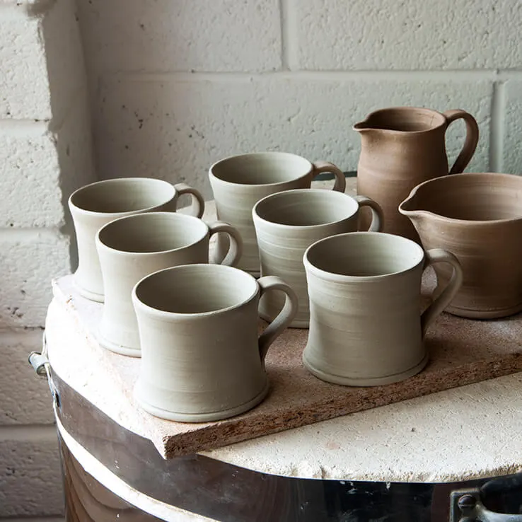 Handmade ceramic mugs and jugs displayed on a wooden shelf against a white brick wall.