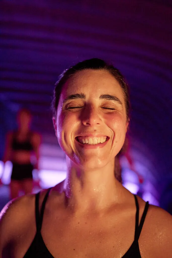 Woman smiling with eyes closed in a purple-lit yoga studio during a class.