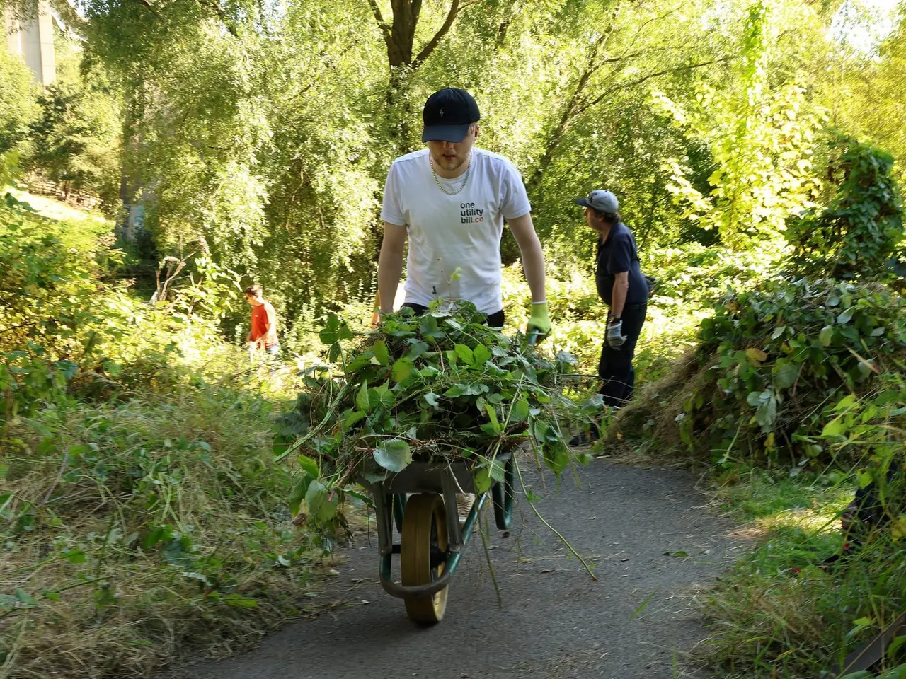 Man pushing a wheelbarrow laden with foliage along a shaded woodland path, with other volunteers nearby.