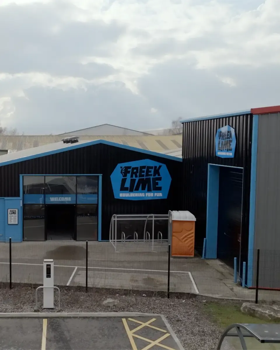 This is a PHOTOGRAPH.

Exterior of Freeklime indoor climbing facility with blue and black corrugated building, entrance area with bike racks and portable toilet visible.