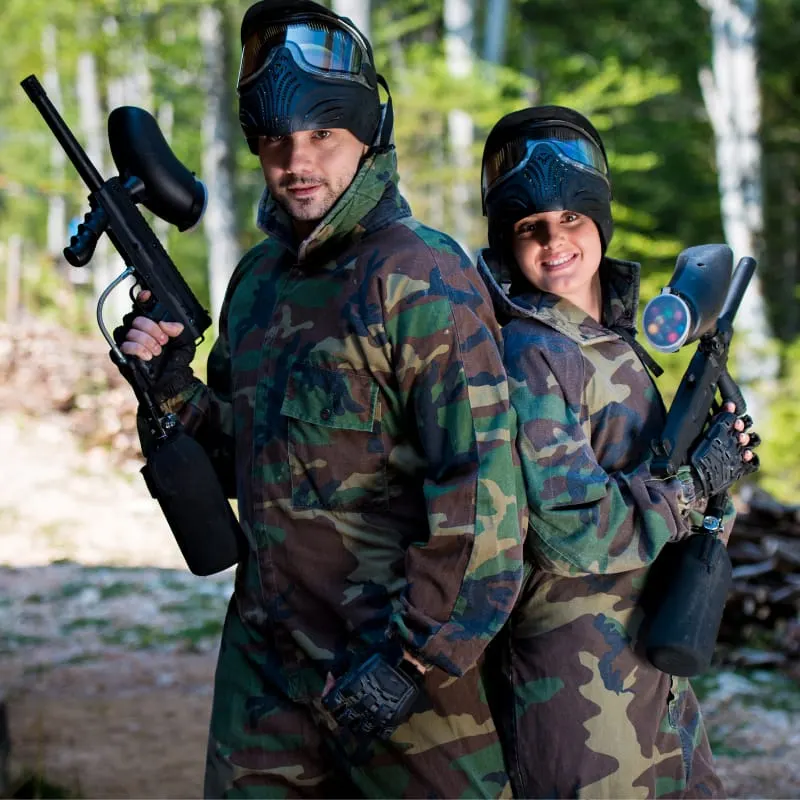 Two people in camouflage clothing and protective masks holding paintball guns in a wooded outdoor setting.